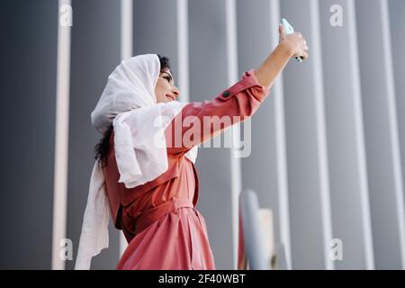 Giovane Araba Donna indossando sciarpa hijab fotografia con uno smartphone all'aperto. Donna araba con hijab che prende selfie con smartphone Foto Stock