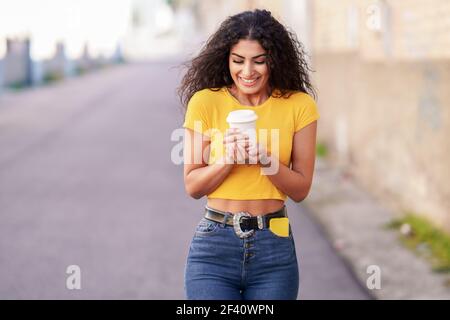 Giovane donna araba che attraversa una strada urbana con un caffè da take-away. Ragazza araba che attraversa la strada con un caffè da take-away Foto Stock