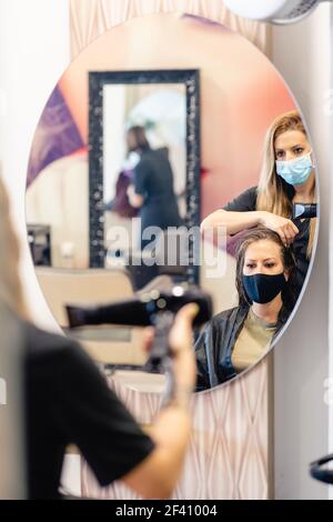 Donna parrucchiera che asciuga i capelli del suo cliente&rsquo;s con un asciugacapelli, indossando maschere protettive in un centro di bellezza. Concetti di affari e bellezza. Parrucchiere che asciuga i capelli del suo cliente&rsquo;s con un asciugacapelli che indossa le maschere protettive in un centro di bellezza. Foto Stock