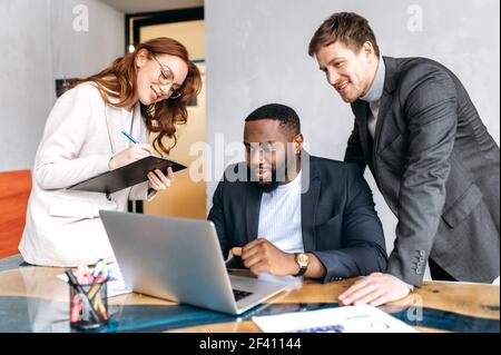 Sorridenti colleghi multietnici stanno lavorando insieme a un nuovo progetto, utilizzando un notebook. Partner commerciali felici di successo che guardano lo schermo del computer, navigare in Internet, seduti in ufficio Foto Stock