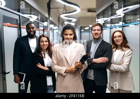 Imprenditori influenti di successo in abiti eleganti sono in piedi in ufficio con mani incrociate. Partner commerciali multirazziali felici che guardano con fiducia alla telecamera, sorridendo Foto Stock