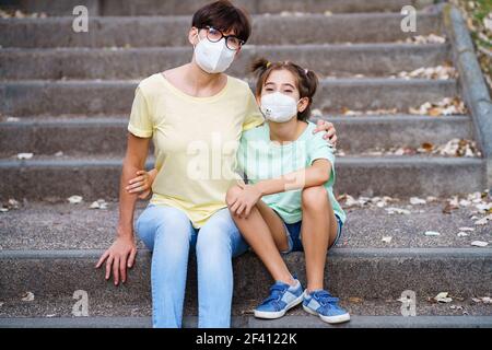 Madre e figlia di mezza età siedono sulla strada indossando maschere a causa della pandemia di Covid-19. Madre e figlia di mezza età siedono sulla strada indossando maschere Foto Stock