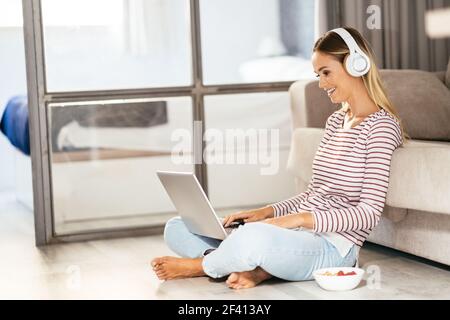 Giovane donna sorridente con cuffie e computer portatile sul divano di casa. Giovane donna sorridente con cuffie e computer portatile sul divano Foto Stock