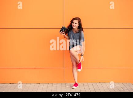 Full Body of trendy girl con fotocamera vintage che mette su sneaker rosse su sfondo arancione. Full Body di ragazza trendy con fotocamera vintage su sfondo arancione Foto Stock