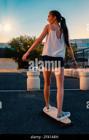 Bella ragazza a cavallo skateboard in corto denim pantaloncini, vista dal retro di fronte al cielo di prima sera. Bella ragazza a cavallo skateboard in corto denim shorts, vista dal retro Foto Stock