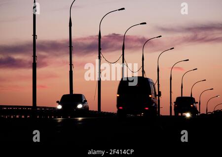 Sagome di auto e furgoni davanti al cielo rosa-arancio del tramonto con sagome di lampioni. Sagome di auto e furgoni davanti al cielo del tramonto con sagome di lampioni Foto Stock