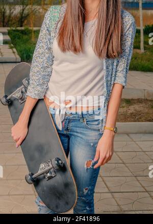 Hipster ragazza in jeans e giacca boho che tiene la vista frontale skateboard. Ragazza hipster in jeans che tengono la vista frontale skateboard Foto Stock