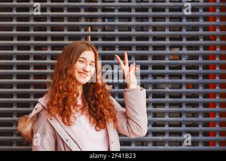 Felice modello rosso mostra V-segno di fronte alla griglia di metallo con un sacco di copyspace. Felice modello rosso mostra V-segno di fronte alla griglia di metallo Foto Stock