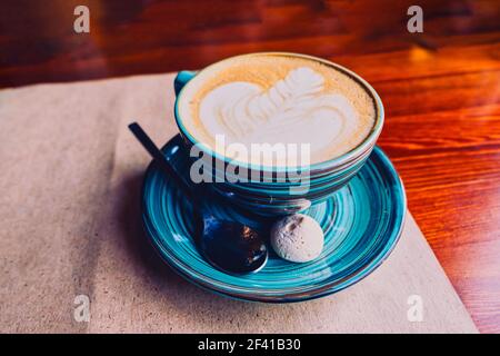 Una grande tazza di cappuccino si trova su un piattino, è servita su un foglio di carta Kraft nella caffetteria. Su un piatto c'è un cucchiaio e una piccola torta di marshmallow. CopySpace è sopra il piatto.. Una tazza verde di cappuccino si trova su un piattino, viene servita su un foglio di carta Kraft nella caffetteria. Su un piatto c'è un cucchiaio e una piccola torta di marshmallow. Foto Stock