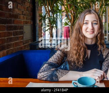 Riposatevi e divertitevi nella caffetteria. Una ragazza sorridente, che rombo guarda direttamente nella macchina fotografica seduto ad un tavolo in un caffè. La parte anteriore è un ragazzino con tè, è vestita di un vestito nero. Una ragazza sorridente, che rombo guarda direttamente nella macchina fotografica seduto ad un tavolo in un caffè. Foto Stock