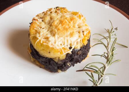 Primo piano di una torta di patate al burro con salsiccia di sangue ricoperta di formaggio fuso grattugiato su un piatto accanto a un fresco rametto di rosmarino. Ricette fatte in casa. Foto Stock
