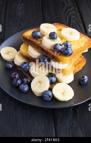 Toast francesi con mirtilli, banana e miele nella piastra nera su fondo di legno scuro. Posizione verticale. Primo piano. Foto Stock