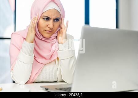 Frustrato lavoratore musulmano di affari overwaged che tocca i templi con le braccia, seduto al portatile, che soffre di forte mal di testa, emicrania, sensazione di ansia, attacco di panico, incapace di affrontare il lavoro Foto Stock