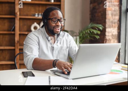 Uomo d'affari afro-americano focalizzato in occhiali indossando cuffie, tenendo videochiamata con i clienti sul laptop, uomo concentrato in occhiali dando online lezione di classe educativa, consultando il cliente Foto Stock