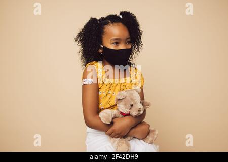 Bambina che indossa una maschera protettiva seduta con il suo orsacchiotto. Bambina con maschera facciale su sfondo marrone dopo aver ricevuto la vaccinazione. Foto Stock