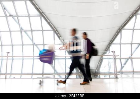 Una gente con carrello che cammina in movimento sfocia su corridoio bianco con grandi finestre. Persone offuscate che camminano in un edificio moderno. Immagine astratta del travel Foto Stock