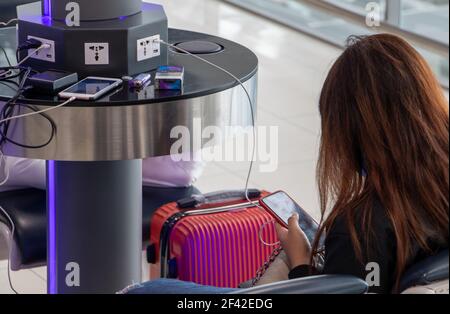 Una giovane donna seduta in corrispondenza di una stazione di carica e guardando il suo smartphone. Ricaricare i telefoni cellulari da libero della stazione di carica in aeroporto. Foto Stock