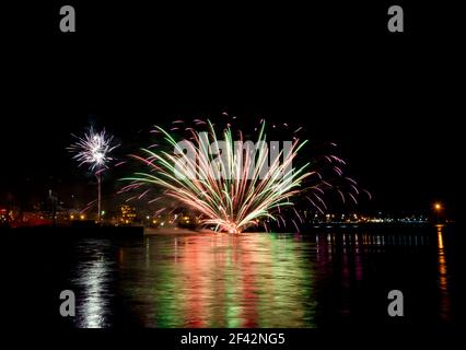 Fuochi d'artificio nautici. Questi esplodono sulla superficie dell'acqua, piuttosto che nell'aria. Gli edifici cittadini sono sullo sfondo. Riflessione sull'acqua. Foto Stock