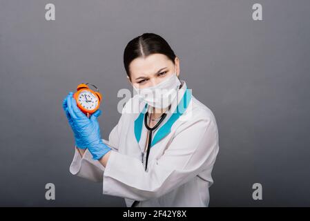 Donna sorridente medico in maschera che punta sull'orologio, tempo di vaccino, concetto medico Foto Stock