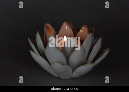 candela da tè illuminata con fiamma in un portacandele in ceramica di fiori di loto su uno sfondo grigio scuro con illuminazione drammatica e romantica e spazio per la copia Foto Stock