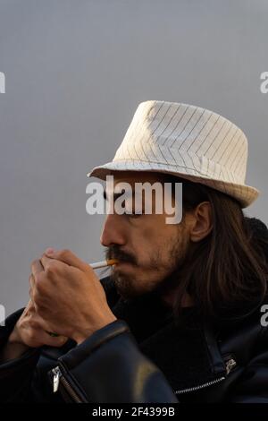 uomo alla moda dai capelli lunghi che fuma sigaretta o tabacco. giovane uomo che illumina sigaretta in giornata ventosa. foto verticale Foto Stock