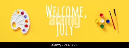 Testo di benvenuto di luglio. Fiori colorati su tavolozza artistica, pennello, gouache su sfondo giallo. Concetto creativo Ciao estate. Banner a disposizione piatta con vista dall'alto. Foto Stock
