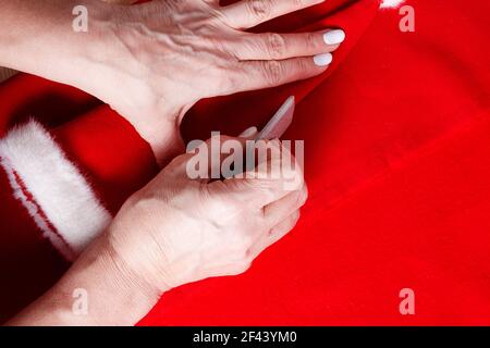 Nonna cucire piccolo cappello di Santa per i nipoti. Primo piano. Gesso del sarto nelle mani per fare il modello di cucitura. Preparazione per la Fiera di Natale della scuola. Foto Stock