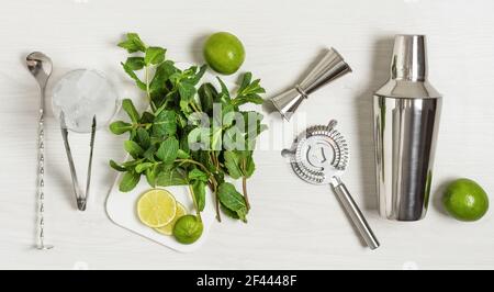 Ingredienti e utensili da bar per preparare un cocktail mojito con cubetti di ghiaccio, menta fresca e lime su sfondo di legno bianco. Set di accessori per barre per moj Foto Stock