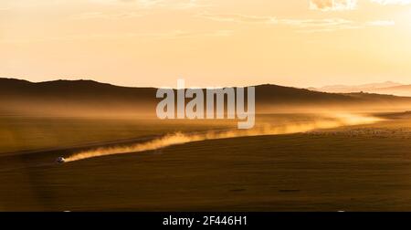 Tramonto sulla steppa della Mongolia con polvere e auto in movimento su una strada sterrata e montagne sullo sfondo. Foto Stock