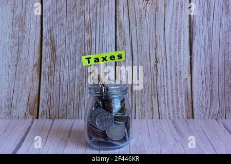 Testo sulla nota gialla su un vaso pieno di monete su una scrivania di legno. Tasse Foto Stock