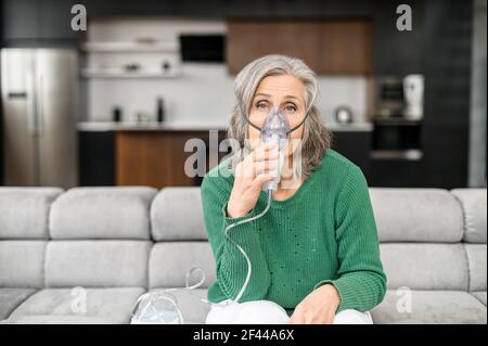 Malata donna anziana con capelli grigi seduta sul divano in un soggiorno, indossando una maschera di ossigeno per trattare l'influenza, la tosse, il coronavirus, facendo l'inalazione attraverso un dispositivo inalatore, sentendosi malato e stanco Foto Stock