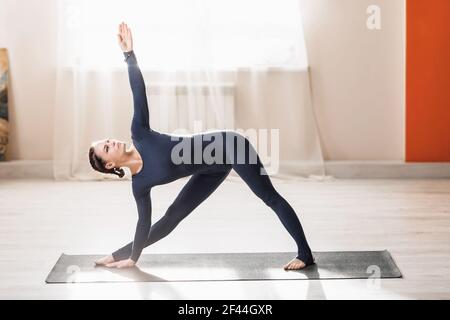 Una bella donna di mezza età che pratica yoga a casa, facendo esercizi triangolari estesi, un allenatore in blu scuro si trova nel Trik Utthita Foto Stock