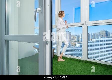 Donna a piedi nudi si erge sul balcone del suo nuovo appartamento e ammira la vista della città dalla finestra Foto Stock