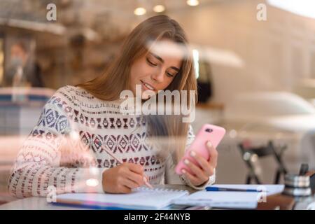 Ragazza affascinante dello studente soddisfatto che usa il telefono cellulare mentre fa i compiti all'interno della caffetteria Foto Stock