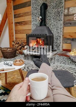 Un primo piano dei piedi di un uomo su una sedia dentro calze in lana grigia allungate verso l'accogliente camino con una tazza di caffè nelle mani Foto Stock