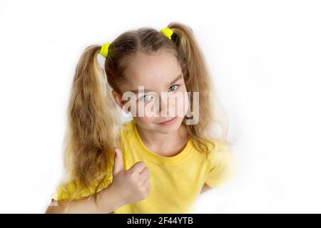 Bambina bionda in una t-shirt gialla fa gesti, alzando le dita, isolata su uno sfondo bianco. Concetto di stile di vita infantile Foto Stock