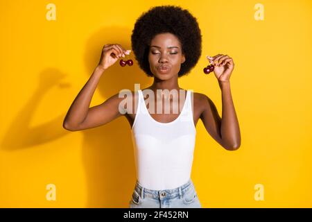 Ritratto di affascinante ragazza dai capelli ondulati e sognante che tiene in mano ciliegie che ti mandano bacio d'aria isolato sul colore giallo brillante sfondo Foto Stock