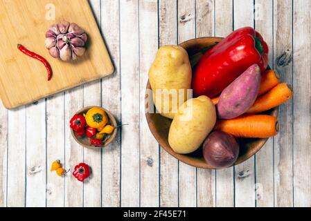 Patate crude, carote e cipolle su una ciotola accanto all'aglio e peperoni su una tavola di legno su un tavolo di legno. Foto Stock