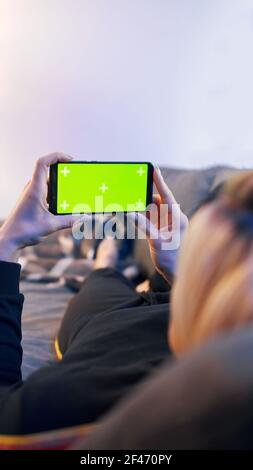 Giovane donna sdraiata su un divano e che utilizza uno smartphone con schermo orizzontale verde. Foto di alta qualità Foto Stock