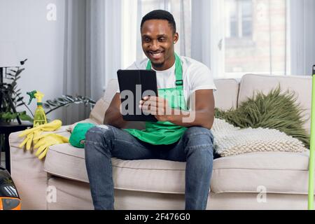 Detergente africano maschile utilizzando tablet digitale mentre si rilassa sul divano e sorridendo facendo una pausa Foto Stock