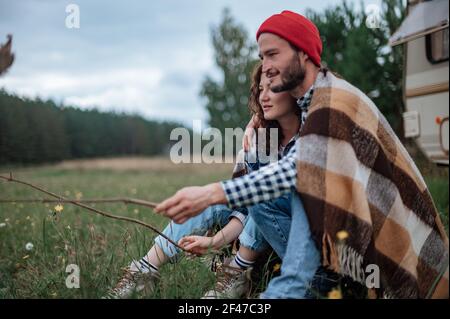 Accoppia in un plaid a scacchi arrostendo marshmallows sul fuoco vicino alla casa del rimorchio. Foto Stock