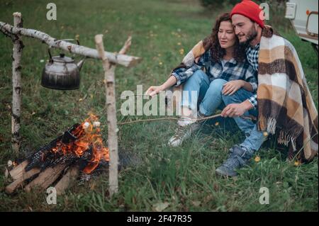 Accoppia in un plaid a scacchi arrostendo marshmallows sul fuoco vicino alla casa del rimorchio. Foto Stock