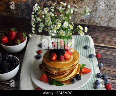 Pancake con frutti di bosco su un piatto bianco. Verticale. Vaso con fiori e frutta piccola. Foto Stock