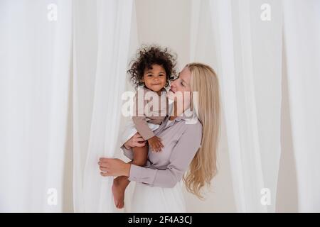 Giovane bionda madre caucasica tiene in braccio la figlia di ricci afroamericana. Donna abbracci ragazza bacia tra i tagli bianchi di tulle fluttuanti luce. Foto Stock