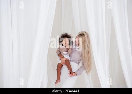 Giovane bionda madre caucasica tiene in braccio la figlia di ricci afroamericana. Donna abbracci ragazza bacia tra i tagli bianchi di tulle fluttuanti luce. Foto Stock