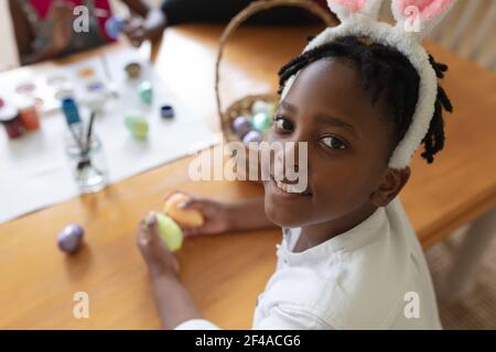 Buon ragazzo afroamericano che indossa orecchie conigliate dipingendo uova colorate Foto Stock