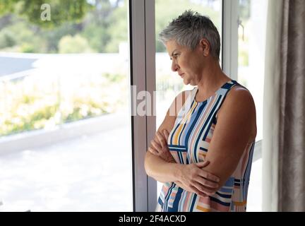 Donna caucasica anziana depressa in piedi guardando fuori dalla finestra di tenuta tazza di caffè Foto Stock