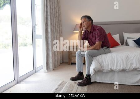 Uomo caucasico anziano seduto sul letto che guarda fuori dalla finestra in camera da letto Foto Stock