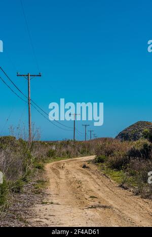 Vecchia strada sterrata di servizio fiancheggiata da pali telefonici e Power Lins sotto un cielo blu. Foto Stock