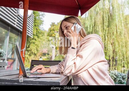 Giovane donna caucasica d'affari con capelli biondi che lavora su un computer portatile in un caffè all'aperto. Studente universitario utilizzando la tecnologia , formazione online, freelance Foto Stock